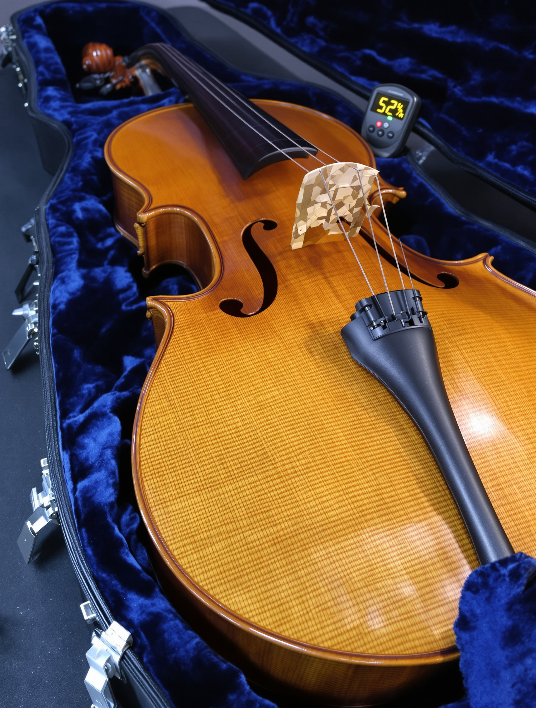 Un violoncelle en érable ondé repose dans son étui rigide ouvert, doublé de velours bleu nuit. Un petit hygromètre numérique placé près de la tête affiche 52%, illustrant le concept de micro-climat protecteur.